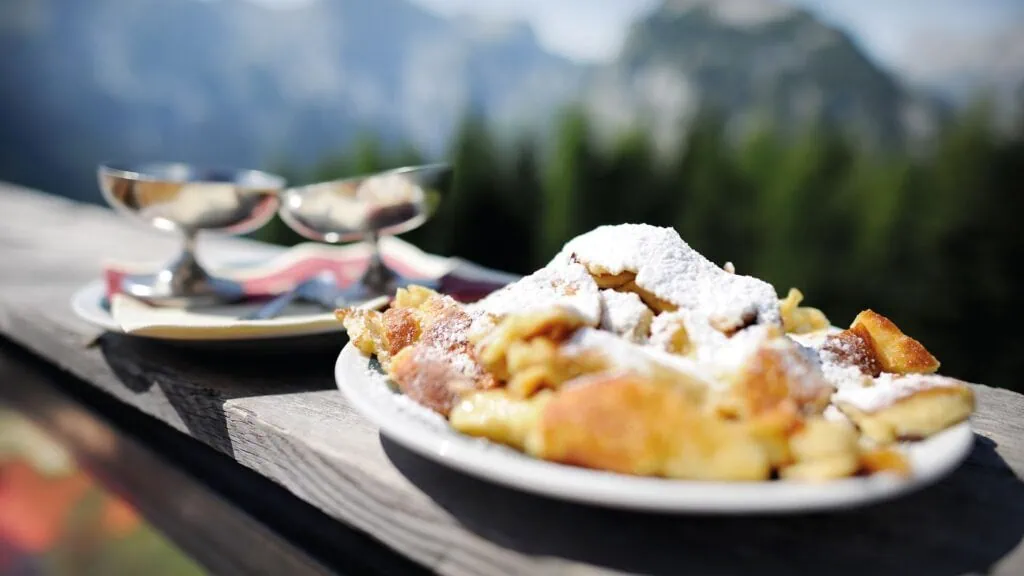 Kaiserschmarrn mit Puderzucker auf Holzbalkon vor Bergpanorama.